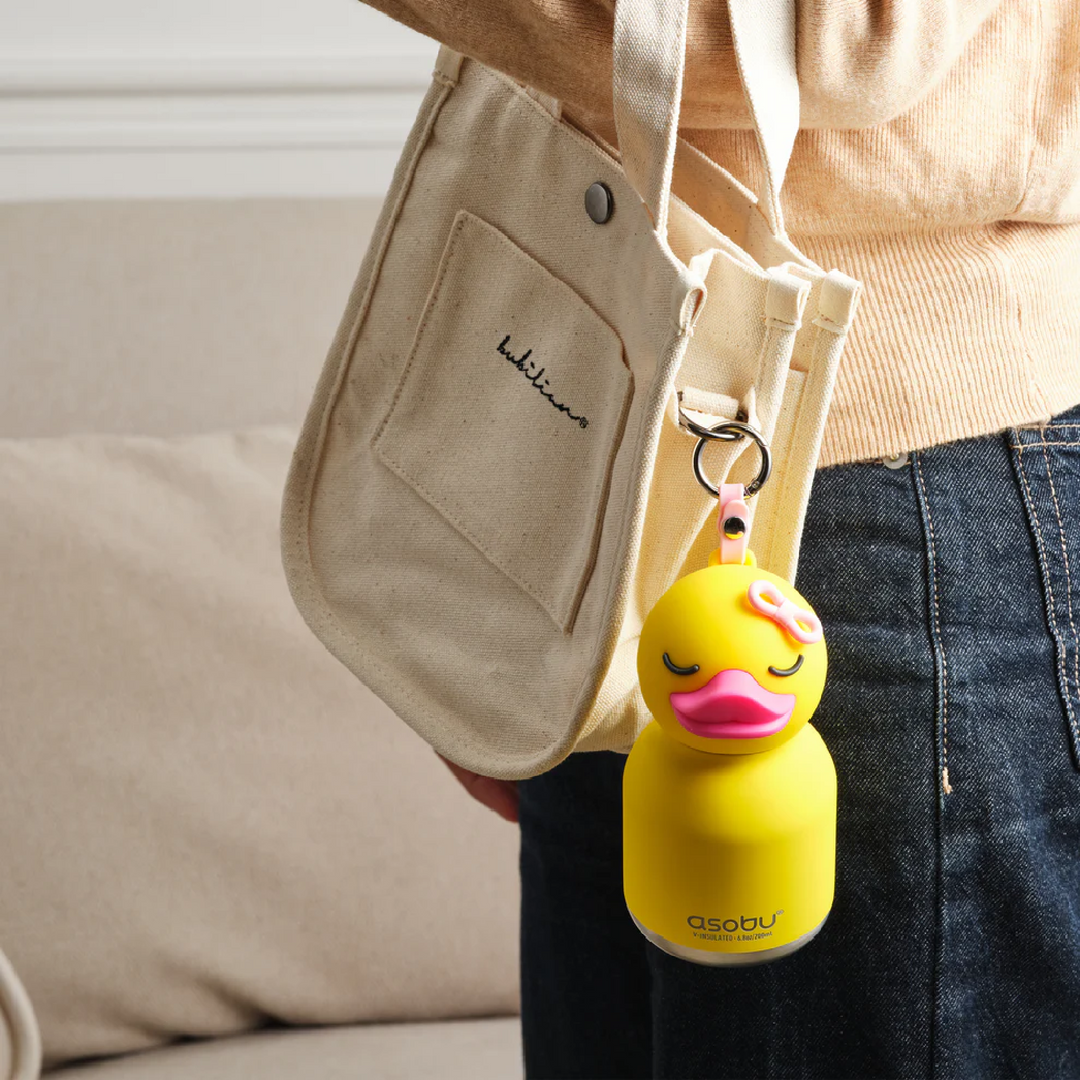 Person holding a beige bag with a yellow duck-shaped bottle holder attached, featuring the brand 'asobu'.